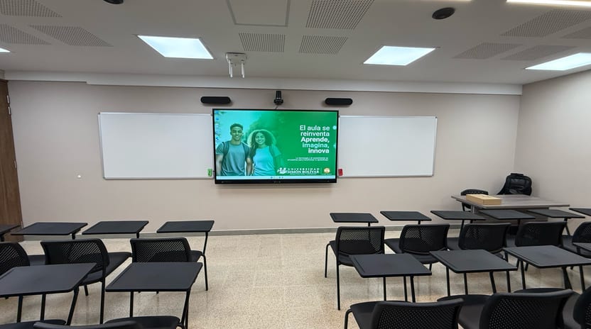 Aula universitaria con la pantalla interactiva al centro del muro, un tablero blanco a cada lado y pupitres frente a ellos