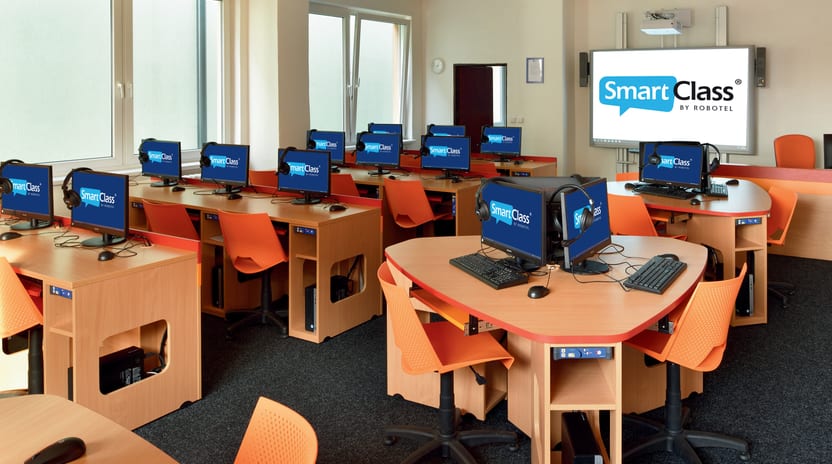 Laboratorio de idiomas con puestos de cómputo en madera, sillas naranja, auriculares en cada puesto y el tablero al frente