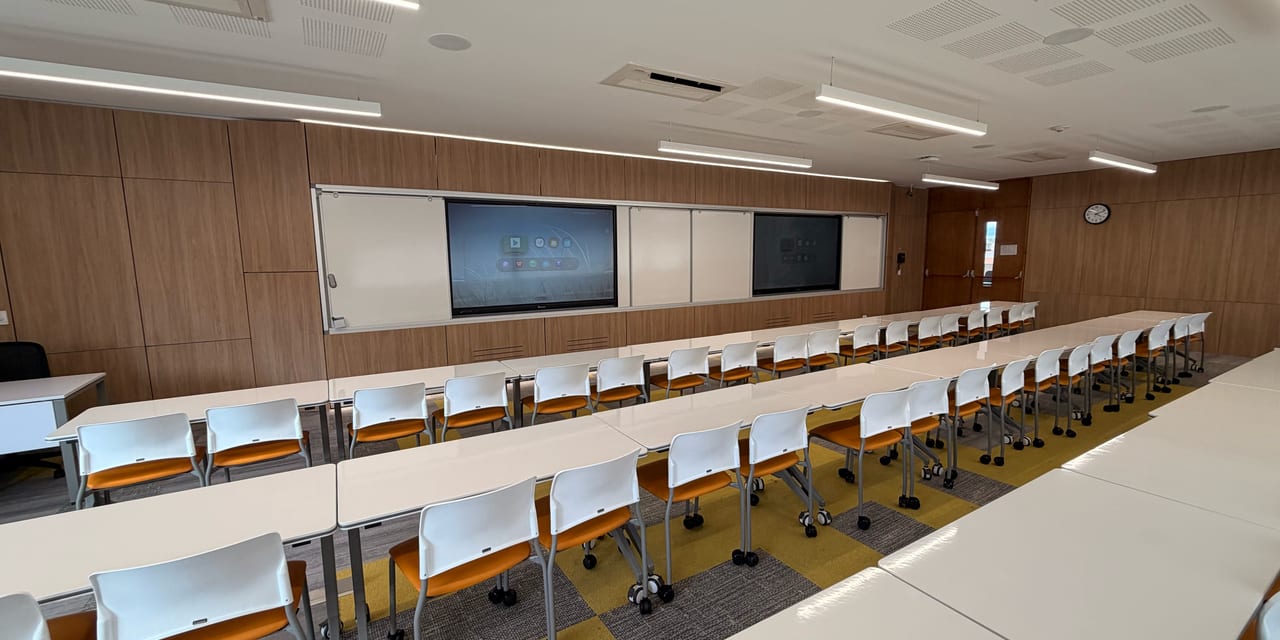 Aula universitaria con dos pantallas interactivas empotradas en un muro de madera, tableros deslizantes y filas de mesas
